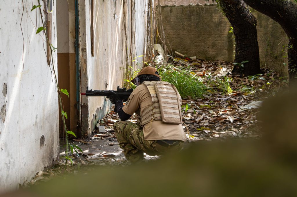 A person in tactical gear engages in airsoft training in an abandoned urban environment.