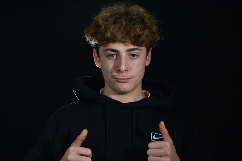 Portrait of a young man in a hoodie with thumbs up, on a dark background.